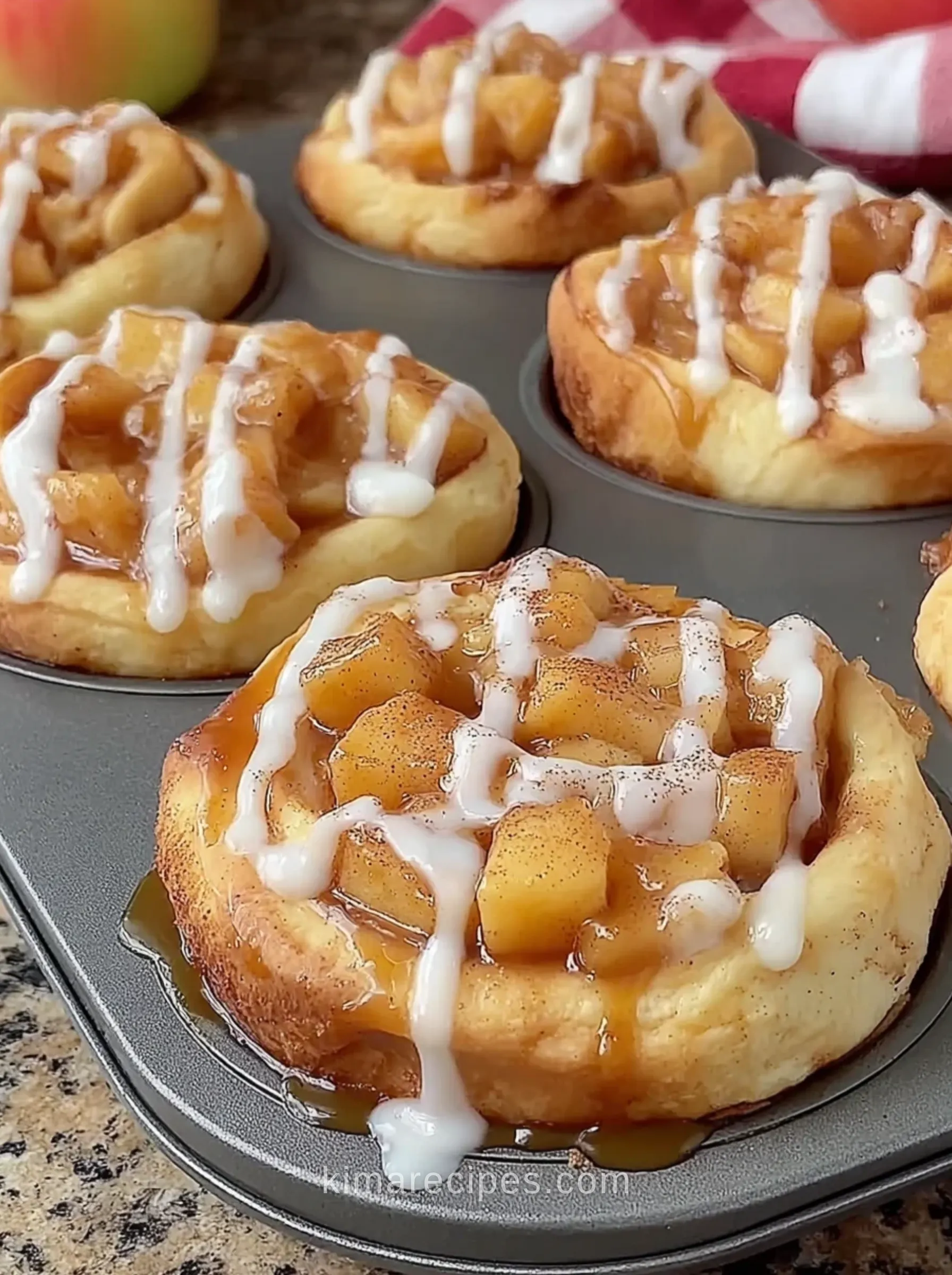 Apple pie cinnamon roll cups in a muffin pan, topped with chopped apple filling and white icing drizzle